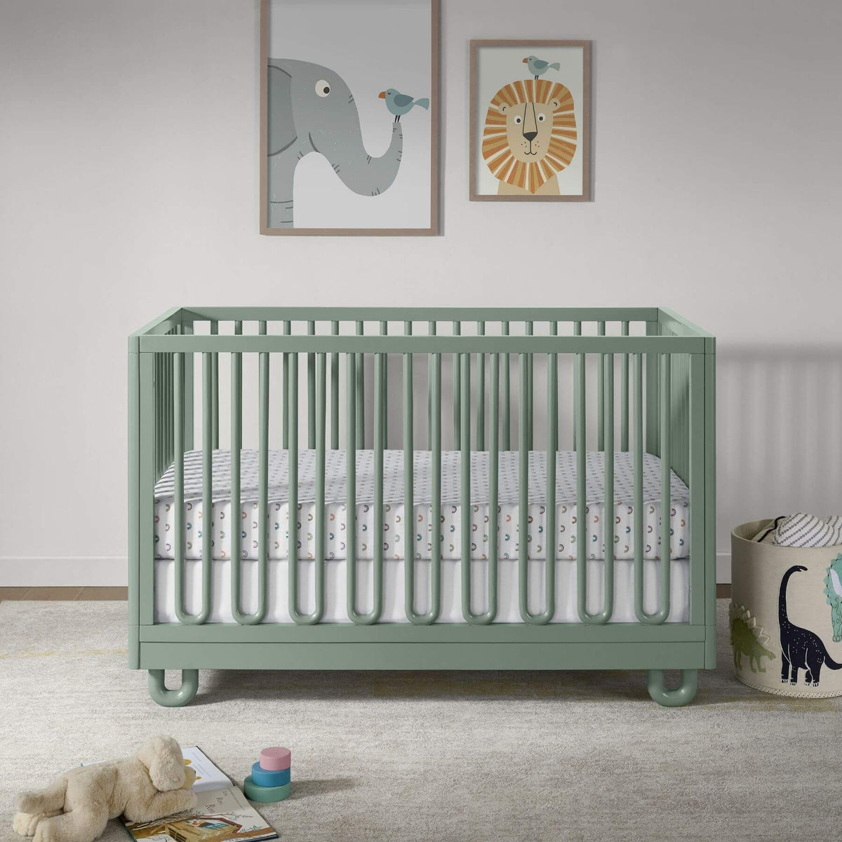 Soho Baby Meridian Island Crib - Olive Green, styled in a nursery with a white mattress, patterned bedding, a stuffed animal, a dinosaur storage bin, and framed animal wall art.