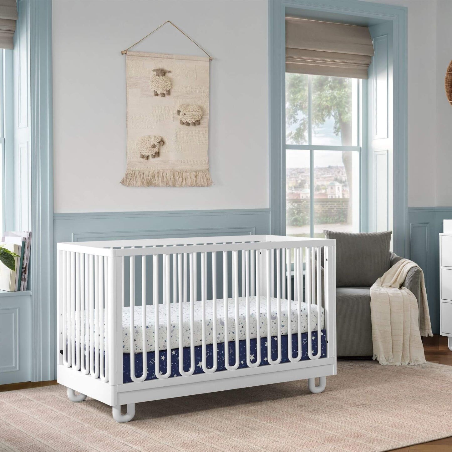 Soho Baby Meridian Island Crib - White with bedding set, styled in a nursery with light blue walls, a sheep wall hanging, and a gray armchair.