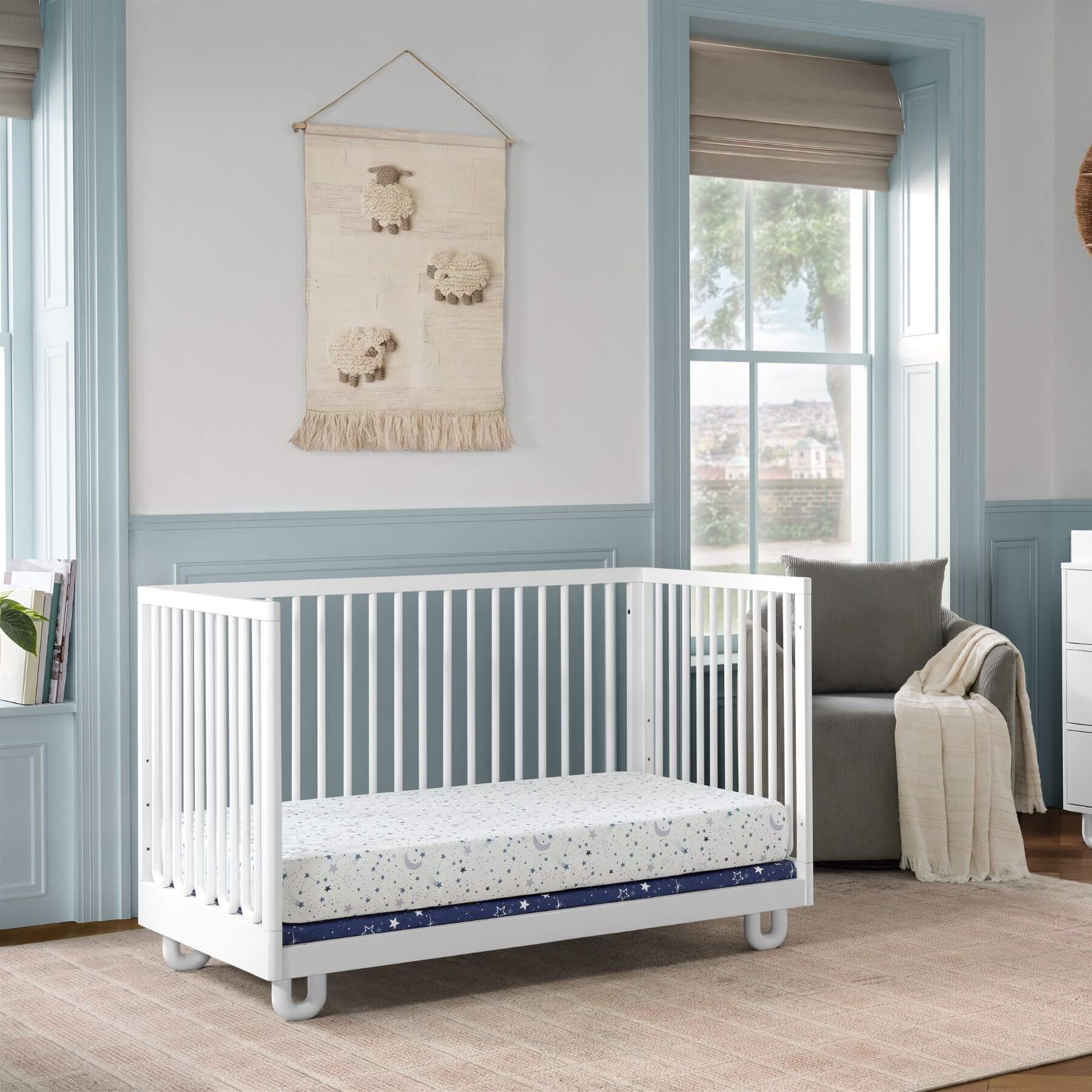 Soho Baby Meridian Island Crib - White and white patterned mattress, styled in a nursery with light blue trim, a sheep wall hanging, and a gray armchair.