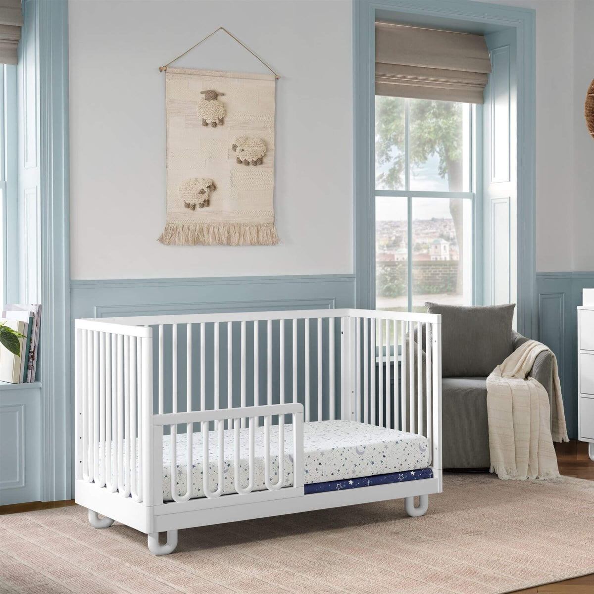 Soho Baby Meridian Island Crib - White with a mattress and patterned sheet in a styled nursery room with blue trim, a window, and a gray armchair.