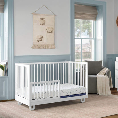 Soho Baby Meridian Island Crib - White with a mattress and patterned sheet in a styled nursery room with blue trim, a window, and a gray armchair.