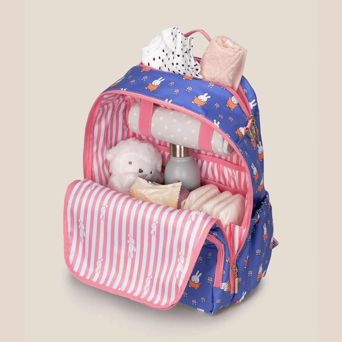 Miffy Flower Fields Zealous Diaper Bag Backpack - Blue & Orange filled with baby essentials, including a white teddy bear, a bottle, and folded cloth diapers, shown on a solid beige background.