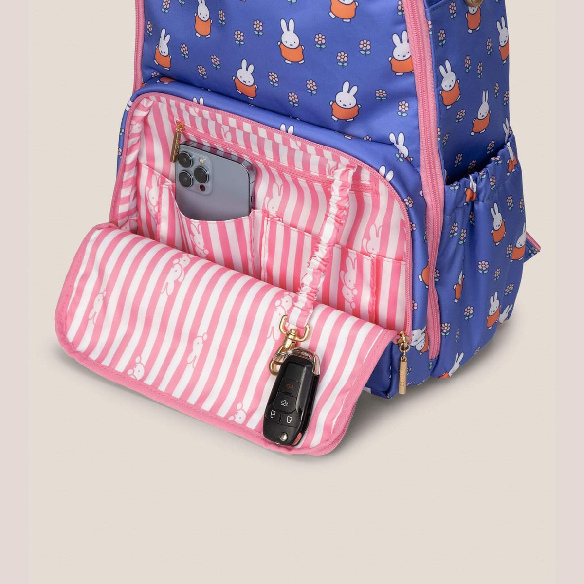 Miffy Flower Fields Zealous Diaper Bag Backpack - Blue & Orange, shown open to reveal a pink-and-white striped interior pocket holding a smartphone and a key fob.