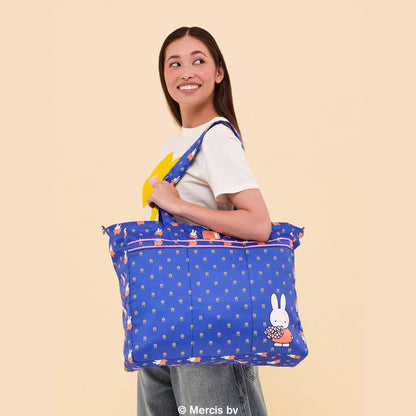A woman wearing a white t-shirt and jeans holds Miffy Flower Fields Super Be large Diaper Bag Duffel - Blue & Orange