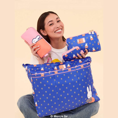 A smiling woman holds several blue Miffy pouches and a large blue bag with a white bunny print, while also holding a pink pouch. She is seated against a plain beige background.