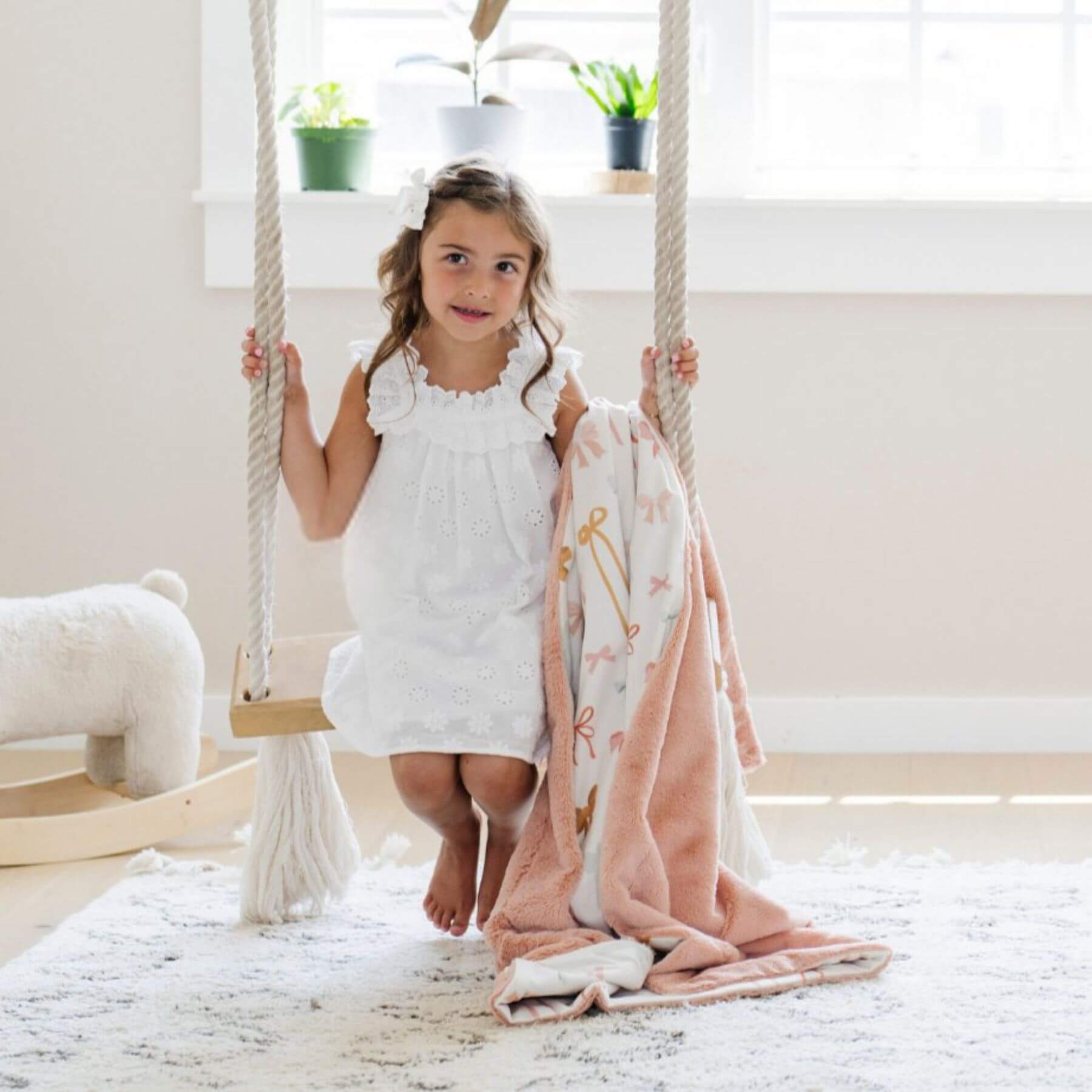 Young girl in a white dress sitting on a wooden swing with Saranoni Minky/Lush Toddler Blanket draped over her, in a bright room with plants.