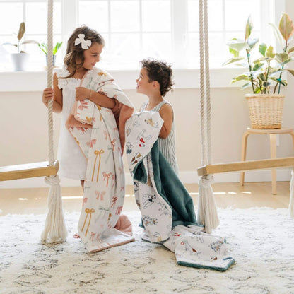 Two children holdingSaranoni Minky/Lush Toddler Blankets on a swing in a bright room with plants.