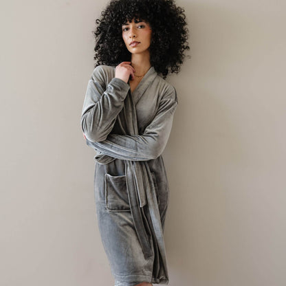 A woman with voluminous curly black hair wearing Saranoni Minky Stretch Luxe Robe - Charcoal, posing against a neutral beige wall.