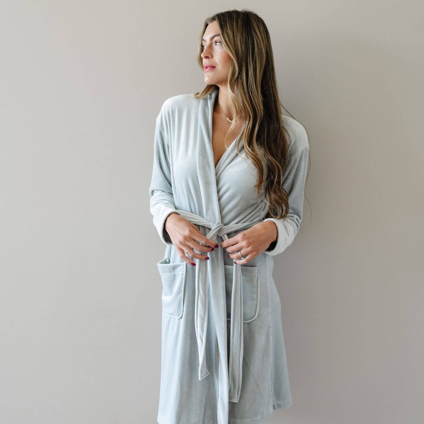 A woman wearing Saranoni Minky Stretch Luxe Robe - Mirage, standing against a plain light gray wall.