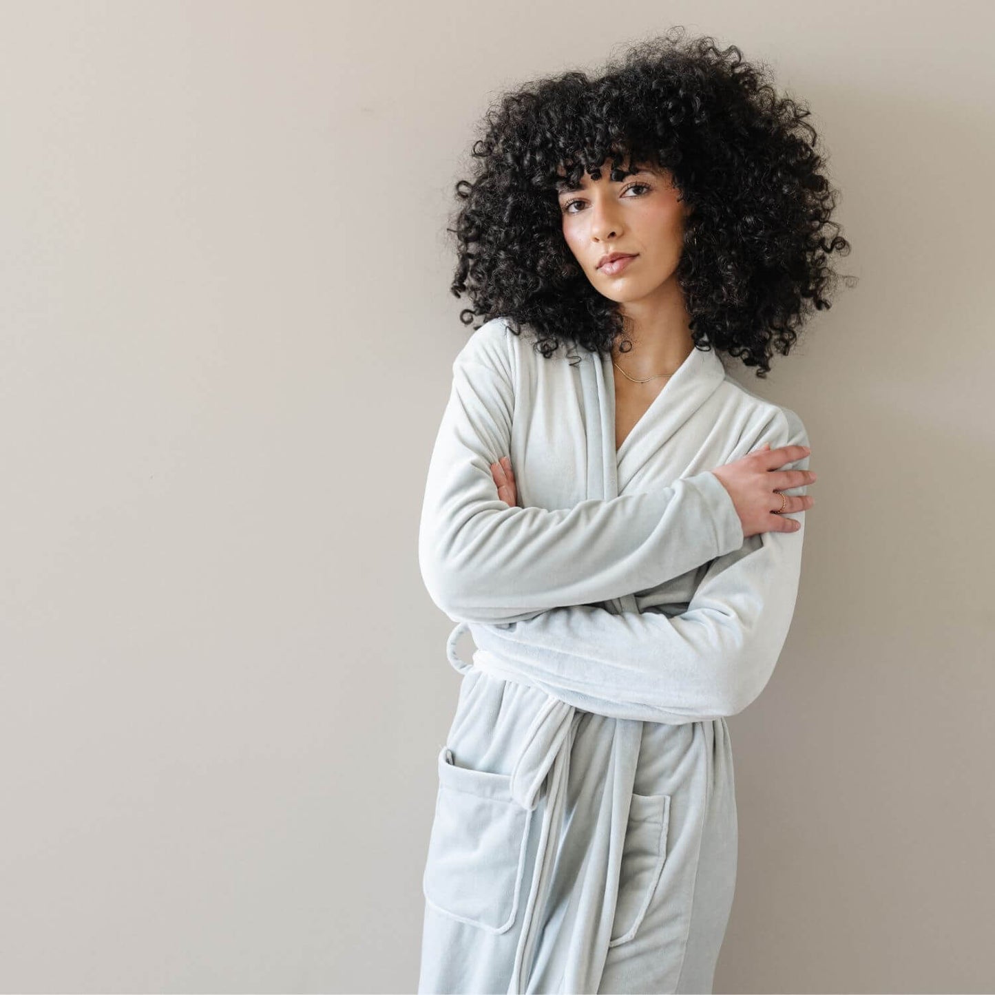 A woman wearing Saranoni Minky Stretch Luxe Robe - Mirage, standing against a plain beige wall with her arms crossed.