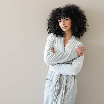 A woman wearing Saranoni Minky Stretch Luxe Robe - Mirage, standing against a plain beige wall with her arms crossed.