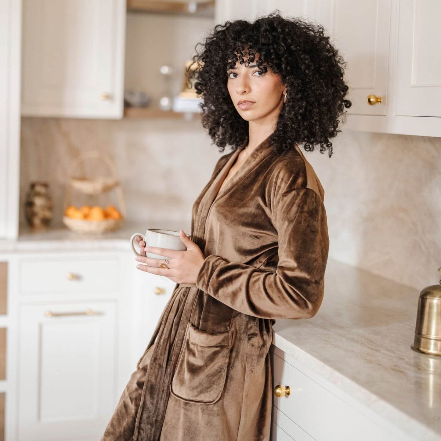 Woman in Saranoni Minky Stretch Luxe Robe - Mocha holding a mug in a kitchen.