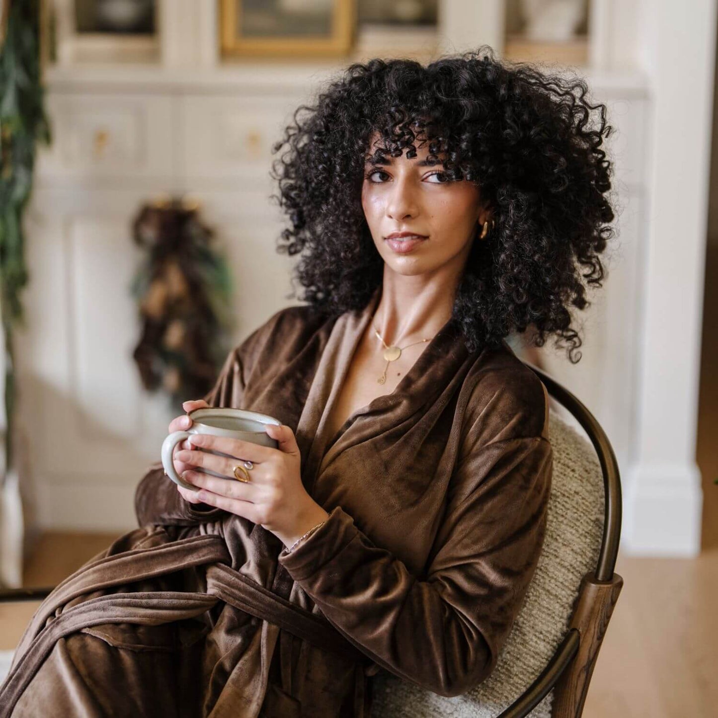 Woman in Saranoni Minky Stretch Luxe Robe - Mocha holding a mug in a cozy room with a fireplace.