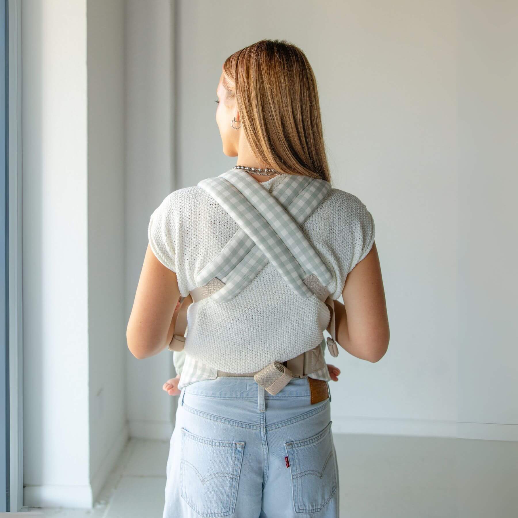 Woman wearing a baby in Monroe x Coco Gingham - Cotton Baby Carrier against a white wall