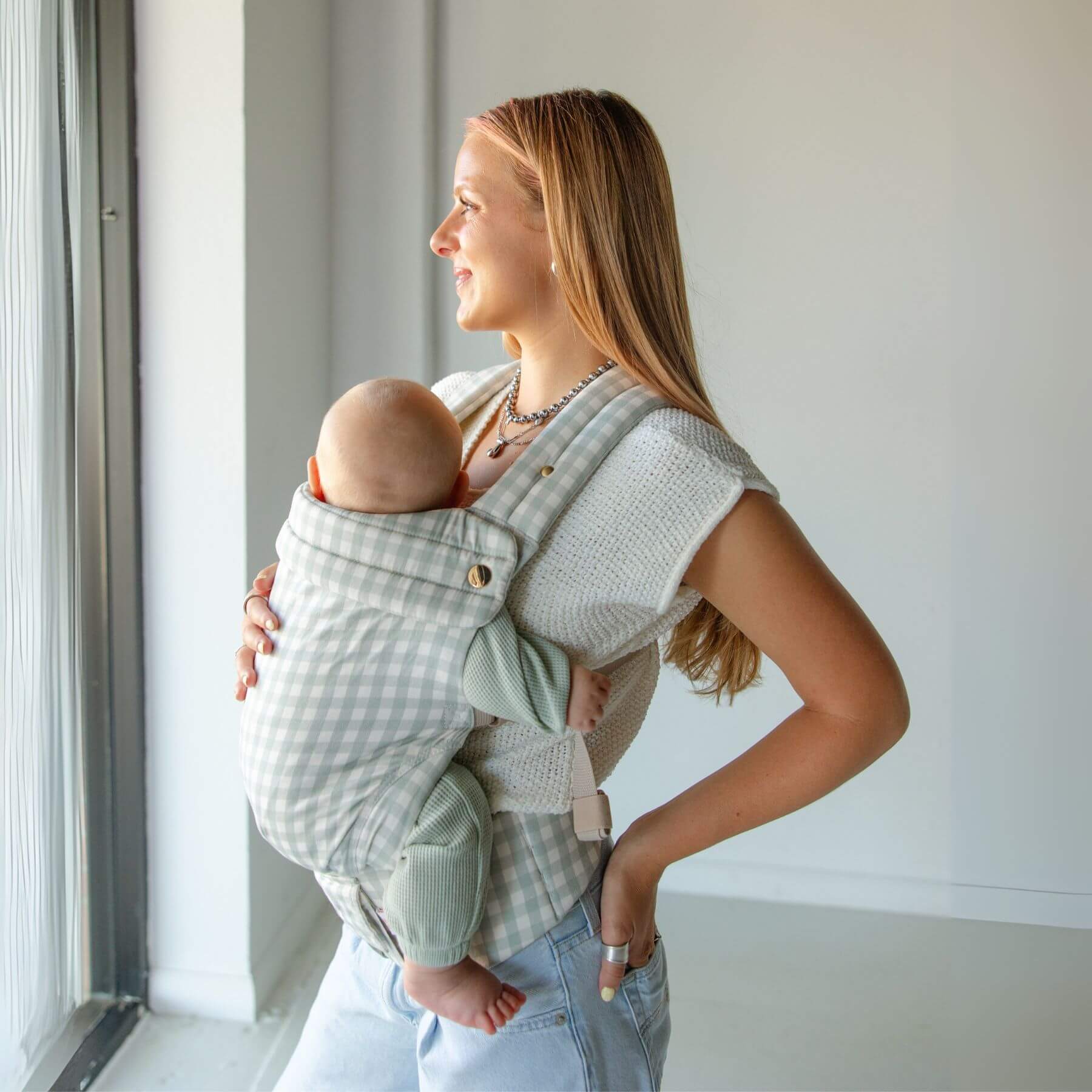 Woman holding a baby in Monroe x Coco Gingham - Cotton Baby Carrier against a white wall.