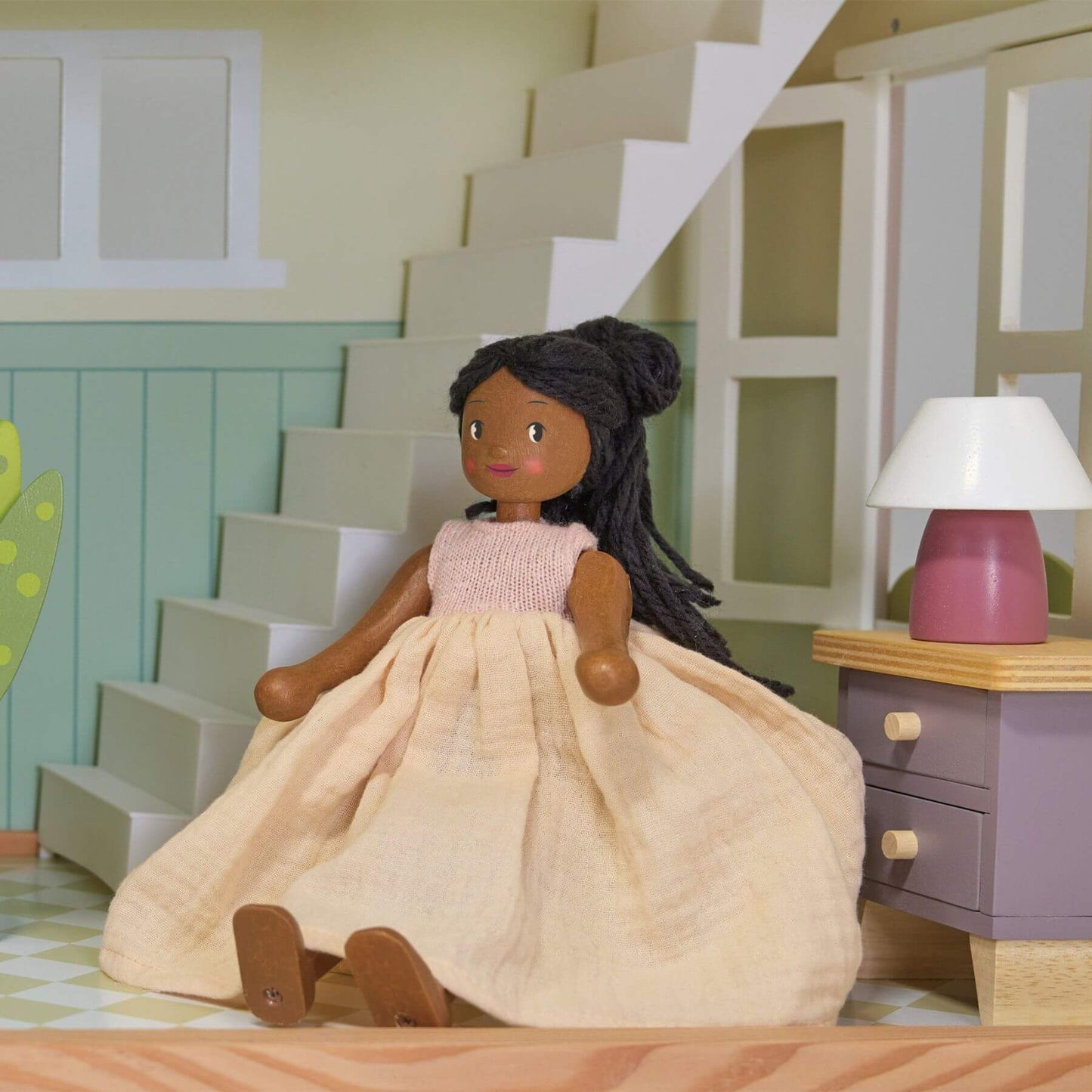 Doll with a beige dress sitting on a wooden floor in a room with a staircase and desk.