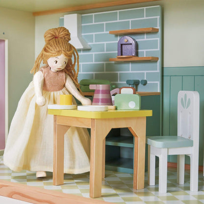 Doll with blonde hair and a white dress sitting at a wooden table in a play kitchen setting.