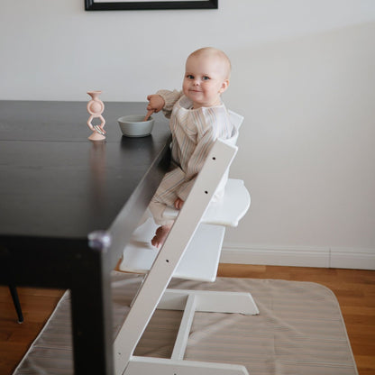 Baby sitting on a white high chair at a dining table with a bowl and spoon.