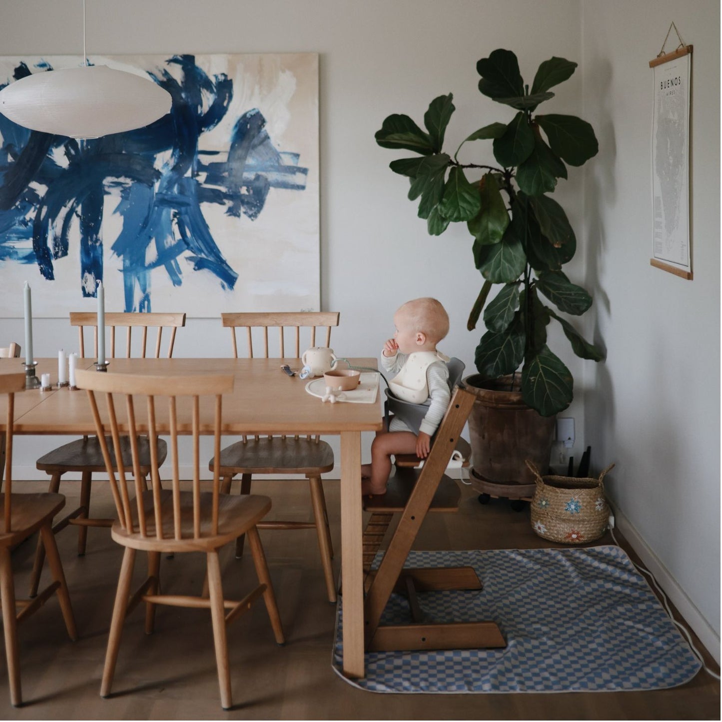 A baby sits in a wooden high chair at a dining table in a bright, modern room with a large blue abstract painting, a potted fiddle-leaf fig plant, and a patterned rug.