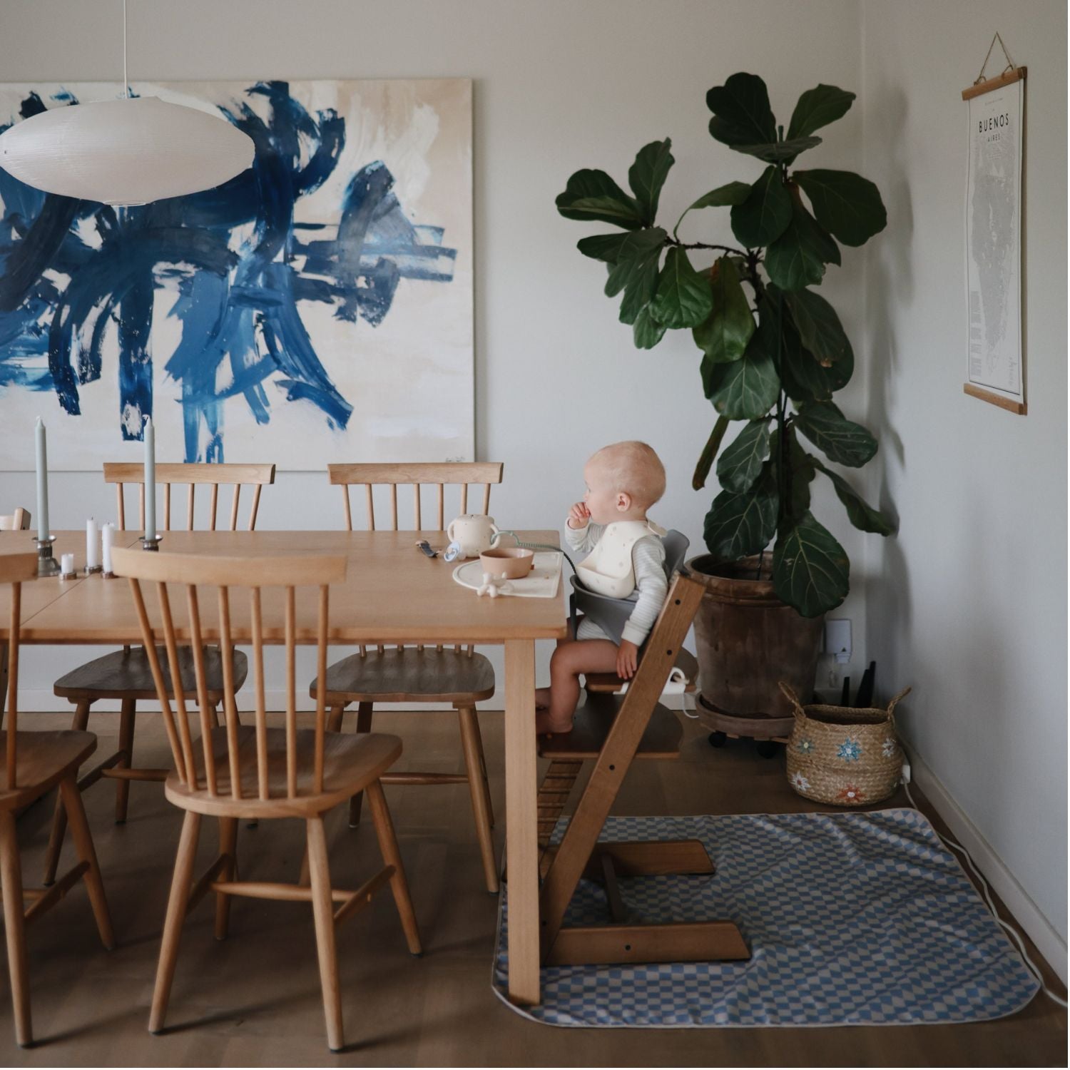 A baby sits in a wooden high chair at a dining table in a bright, modern room with a large blue abstract painting, a potted fiddle-leaf fig plant, and a patterned rug.