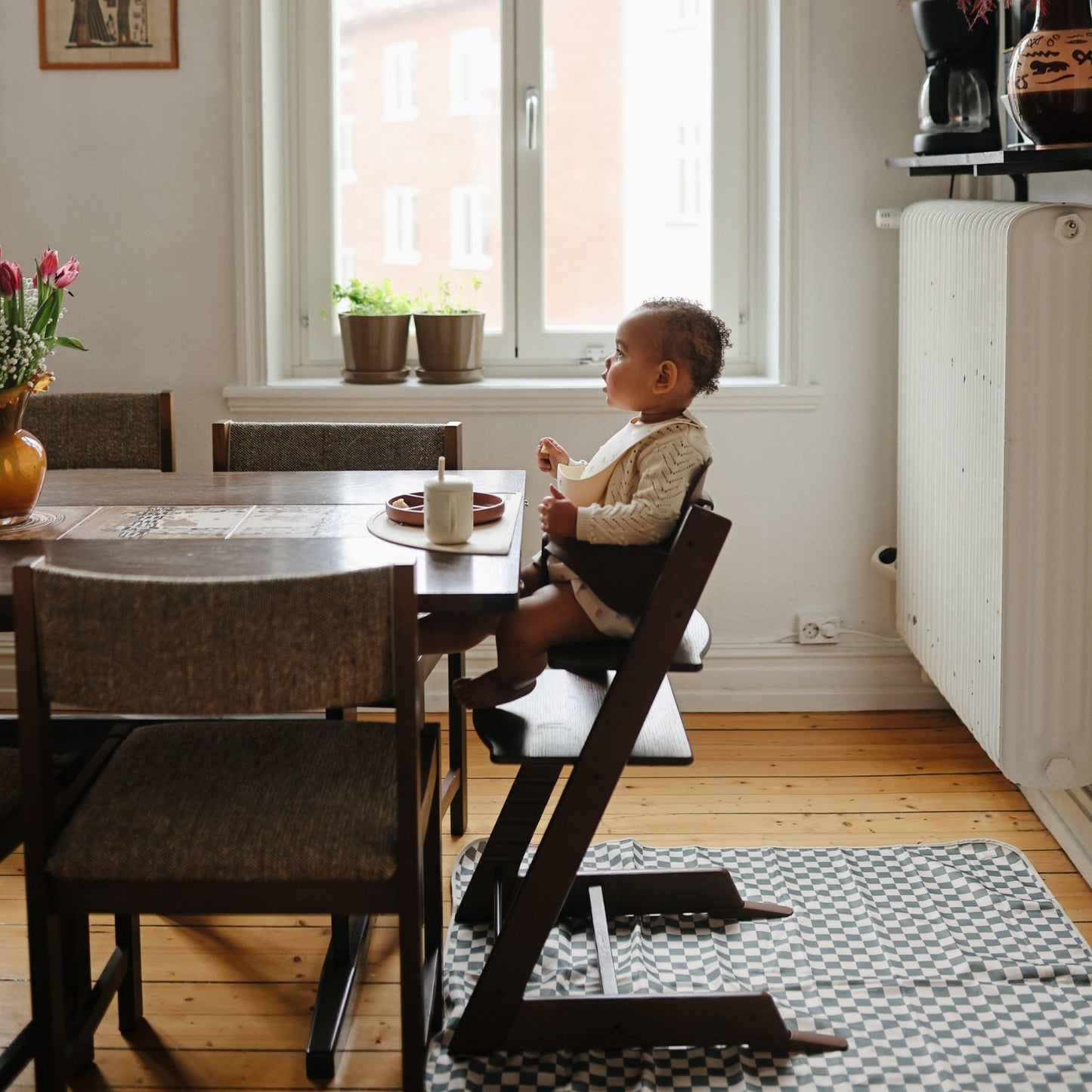 A toddler sits in a dark wood high chair at a dining table in a bright, modern room with a window, radiator, and checkered rug.