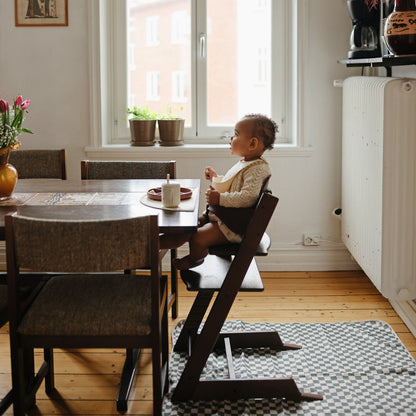 A toddler sits in a dark wood high chair at a dining table in a bright, modern room with a window, radiator, and checkered rug.