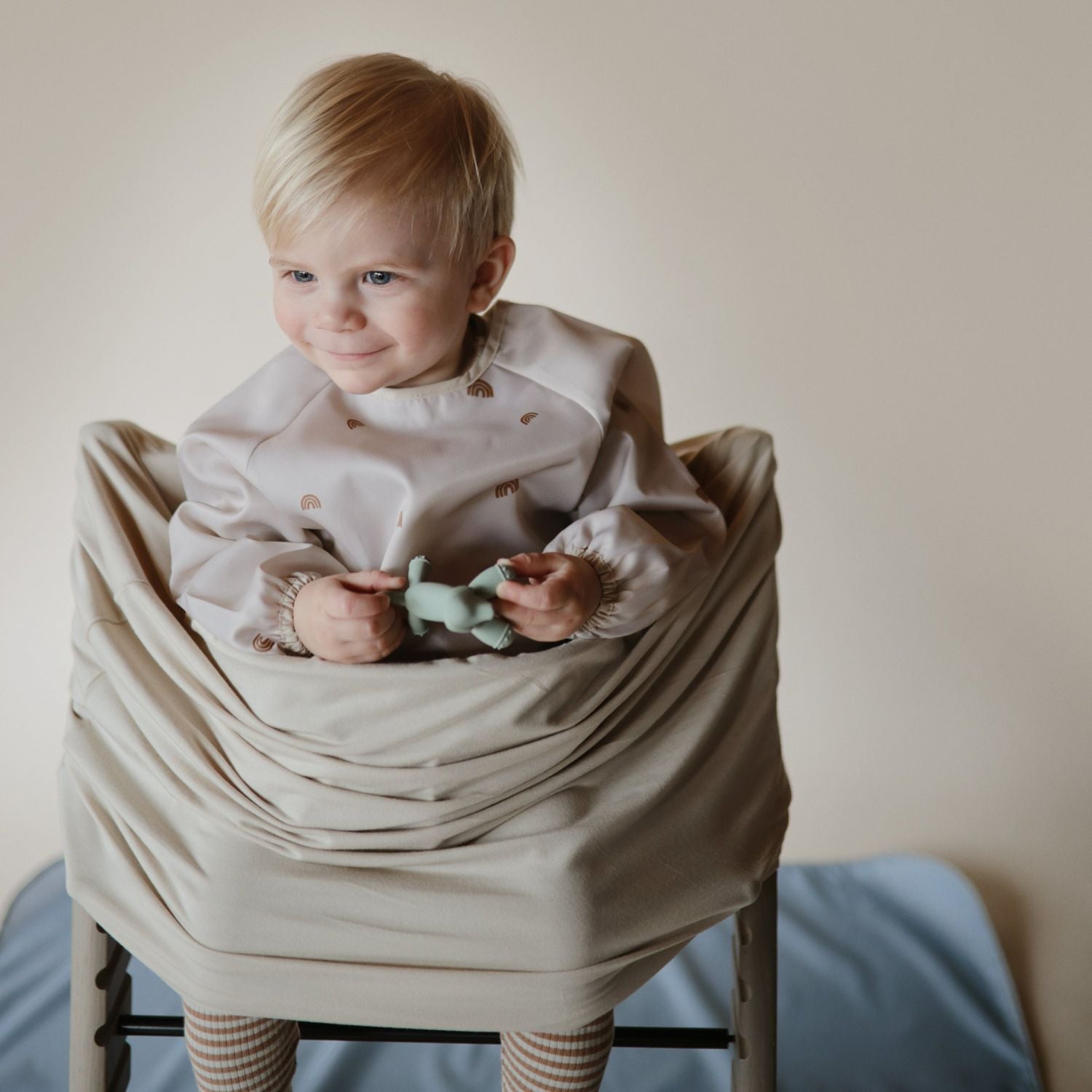 Child sitting on a chair wrapped in a beige blanket holding a green toy.