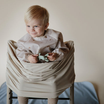 Child sitting on a chair wrapped in a beige blanket holding a green toy.