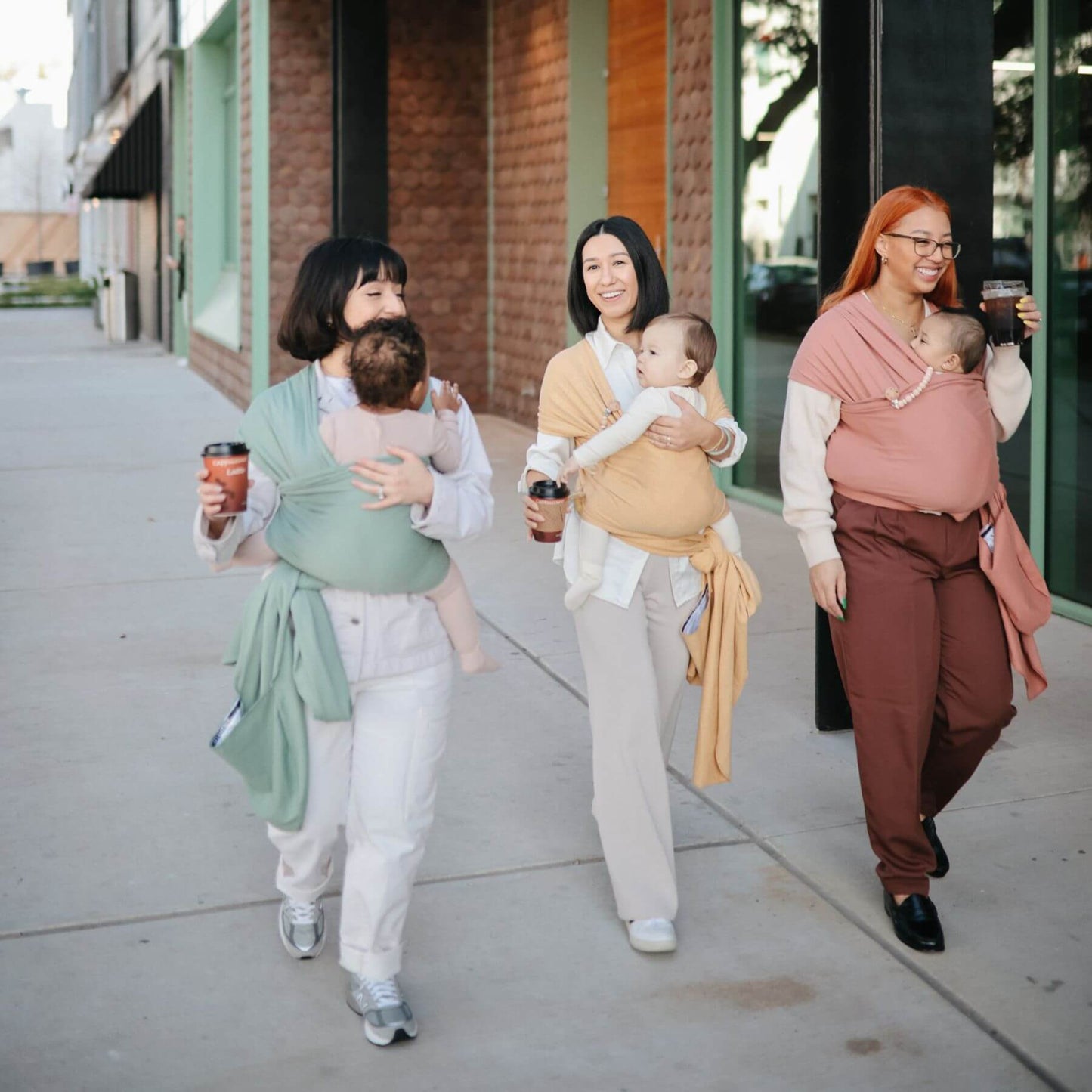 Three women walking on a sidewalk, each carrying a baby in Mushie Baby Wrap.