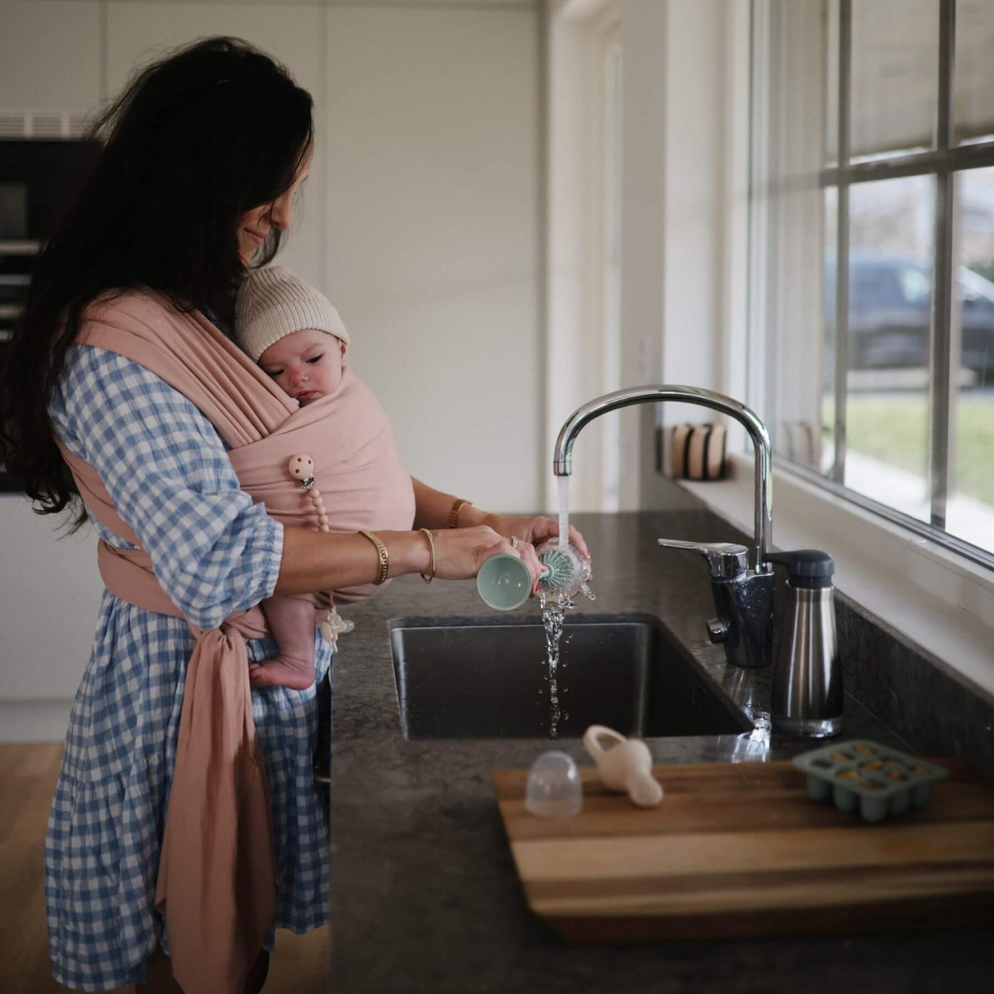 Woman washing dishes in a kitchen with a baby in Mushie Baby Wrap