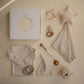 Collection of baby items including a onesie, hat, wrap, and toys on a beige background