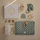 Collection of baby products including a teething blanket, toys, wrap, and a box on a beige background