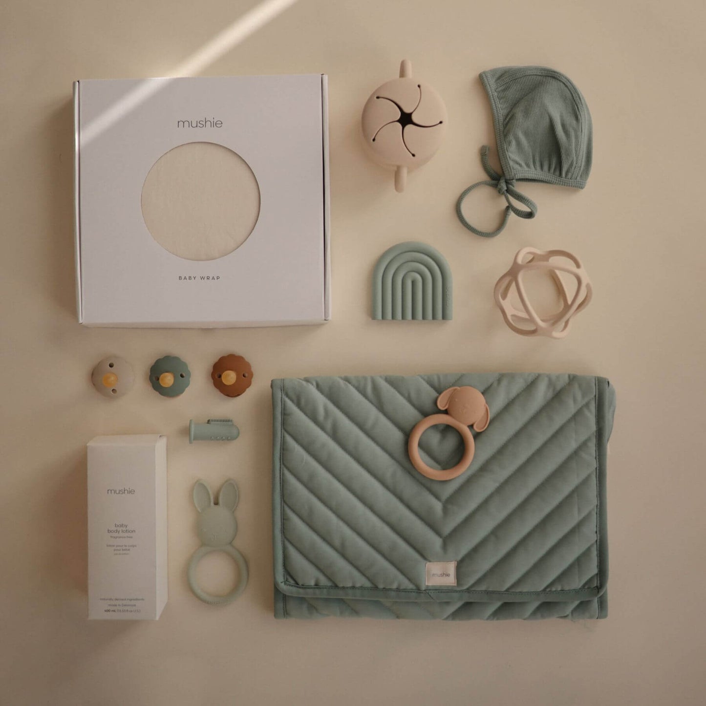 Collection of baby products including a teething blanket, toys, wrap, and a box on a beige background
