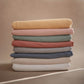 Stack of folded Mushie Baby Wrap in various colors on a beige background