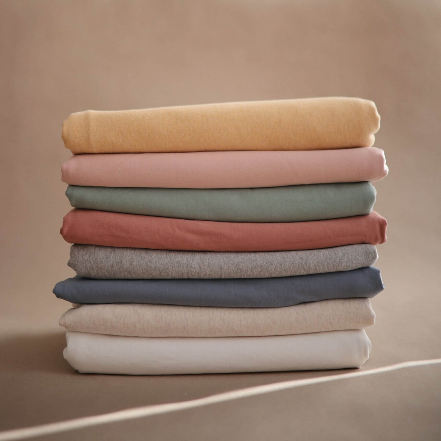 Stack of folded Mushie Baby Wrap in various colors on a beige background