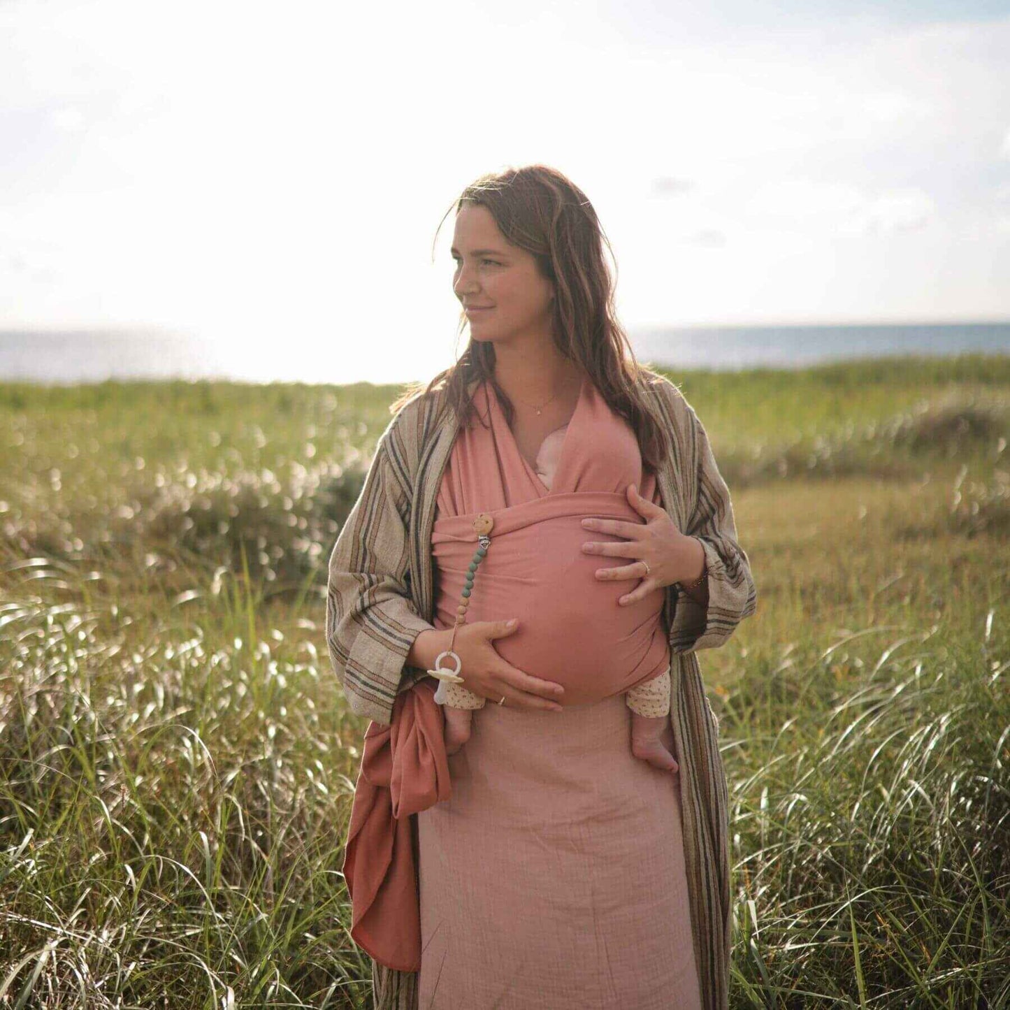 Woman in a pink dress holding a baby Mushie Baby Wrap against a grassy field background
