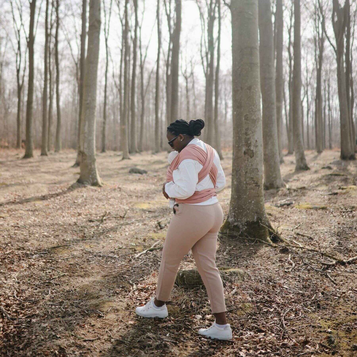 Person standing in a forest holding a baby in Mushie Baby Wrap and beige pants.