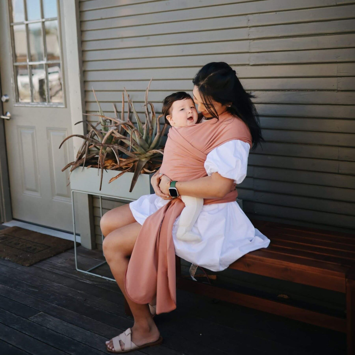Woman sitting on a bench holding a baby wrapped in Mushie Baby Wrap, with a plant and door in the background.