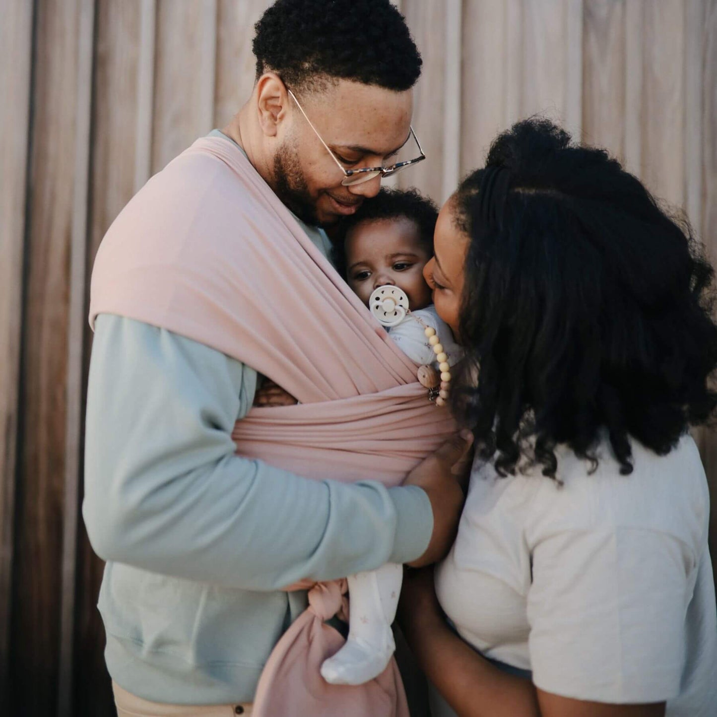 Family with a baby in Mushie Baby Wrap, standing close together outdoors.