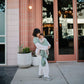 Woman holding a child in Mushie Baby Wrap outside a building with pink doors and white planters.