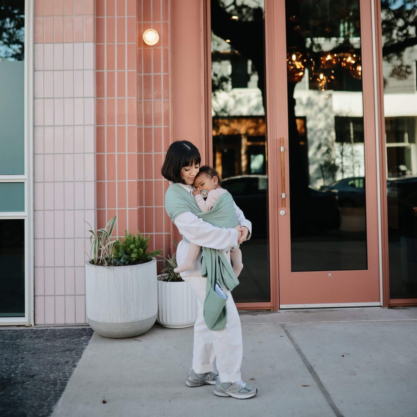 Woman holding a child in Mushie Baby Wrap outside a building with pink doors and white planters.