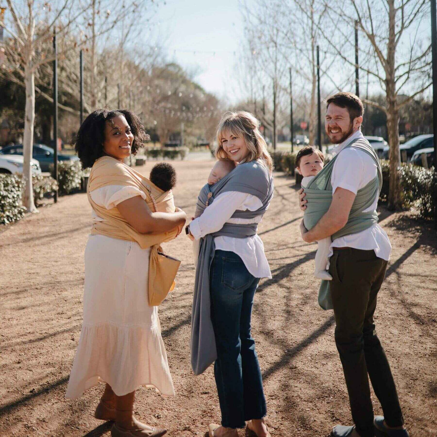 Three adults walking outdoors with children in Mushie Baby Wraps on a sunny day.