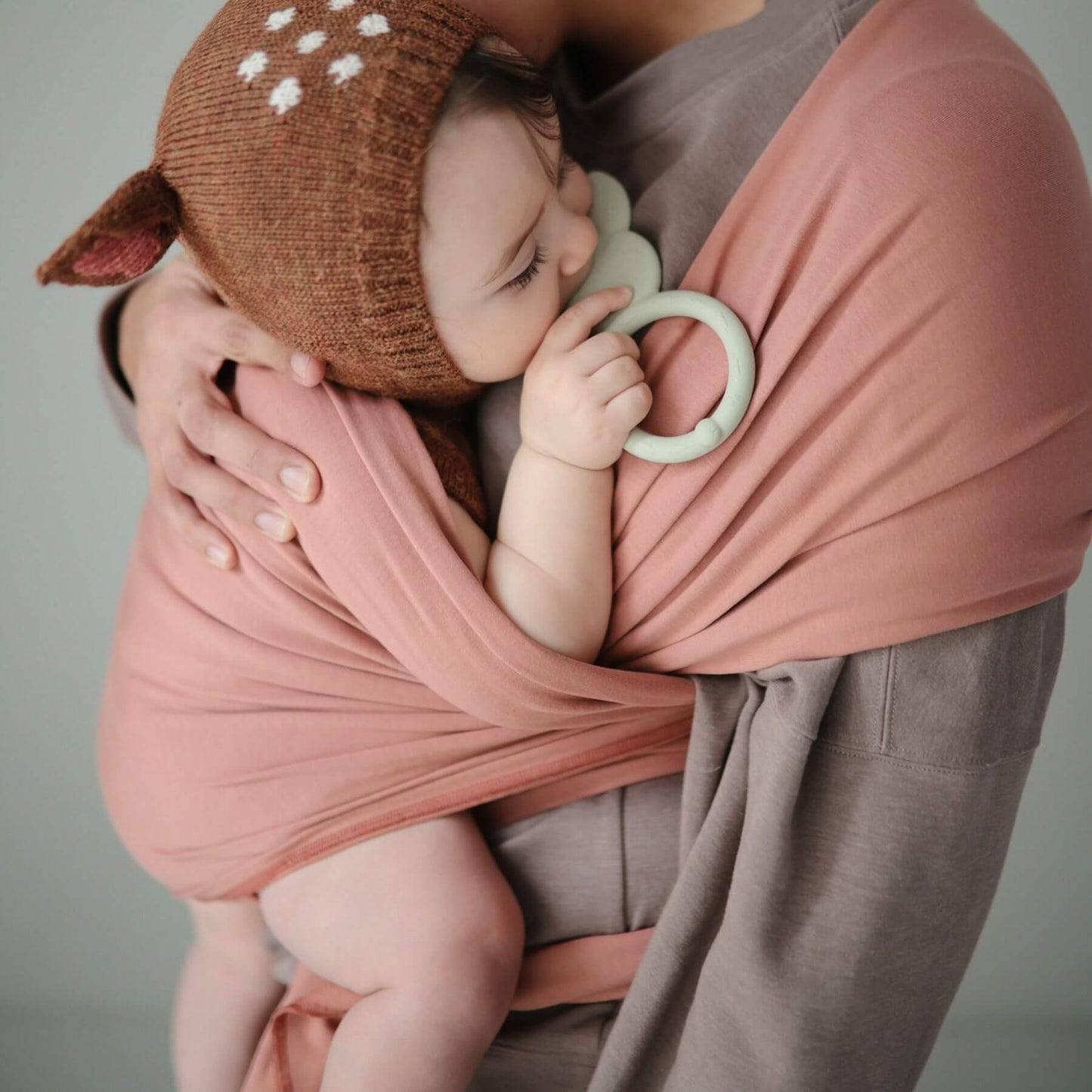 Baby in Mushie Baby Wrap wearing a brown knit hat with deer ears, holding a green teething ring.