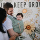 Man holding a baby in a Mushie Baby Wrap with a floral background and text 'Just Keep Growing'.