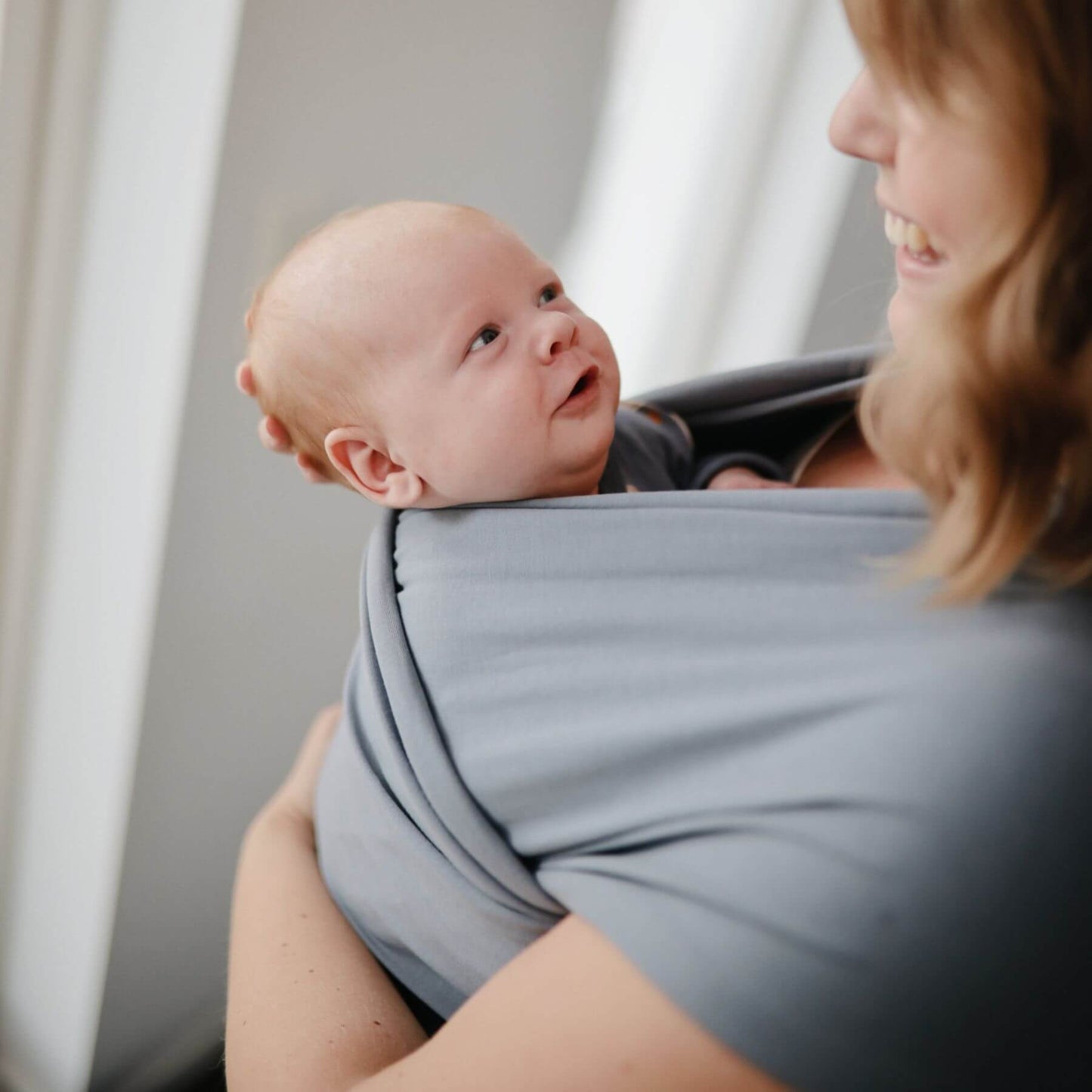 Woman holding a baby wrapped in Mushie Baby Wrap against a neutral background