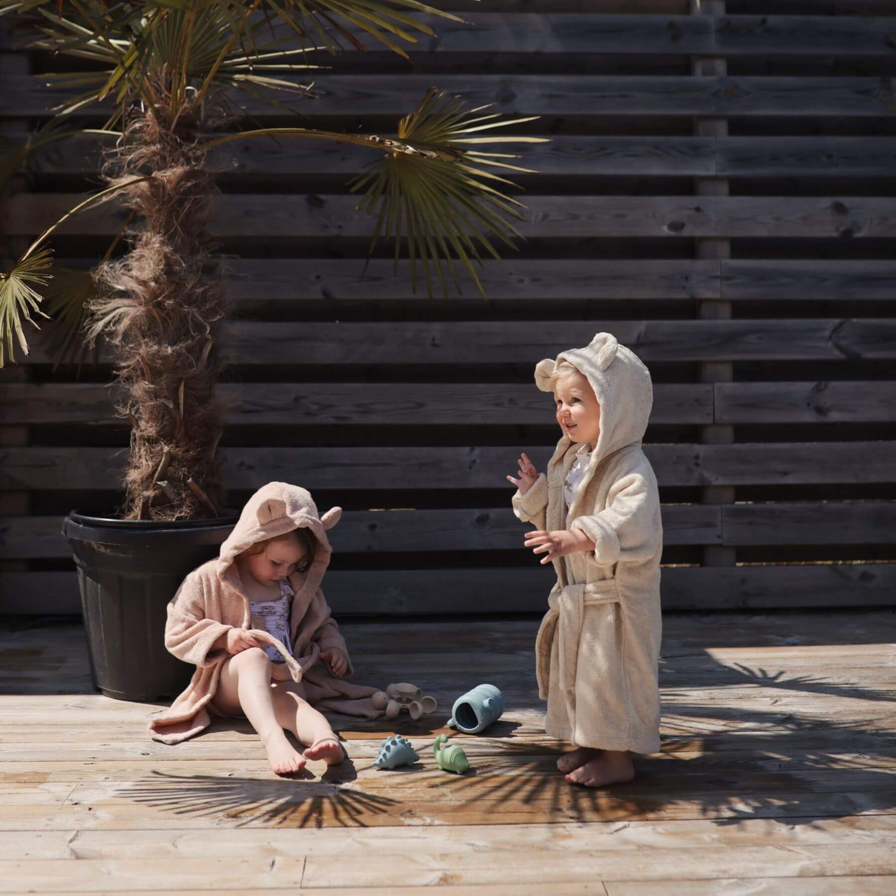 Two children in Mushie Bear Robe playing outdoors on a wooden deck with a wooden wall and plant in the background.