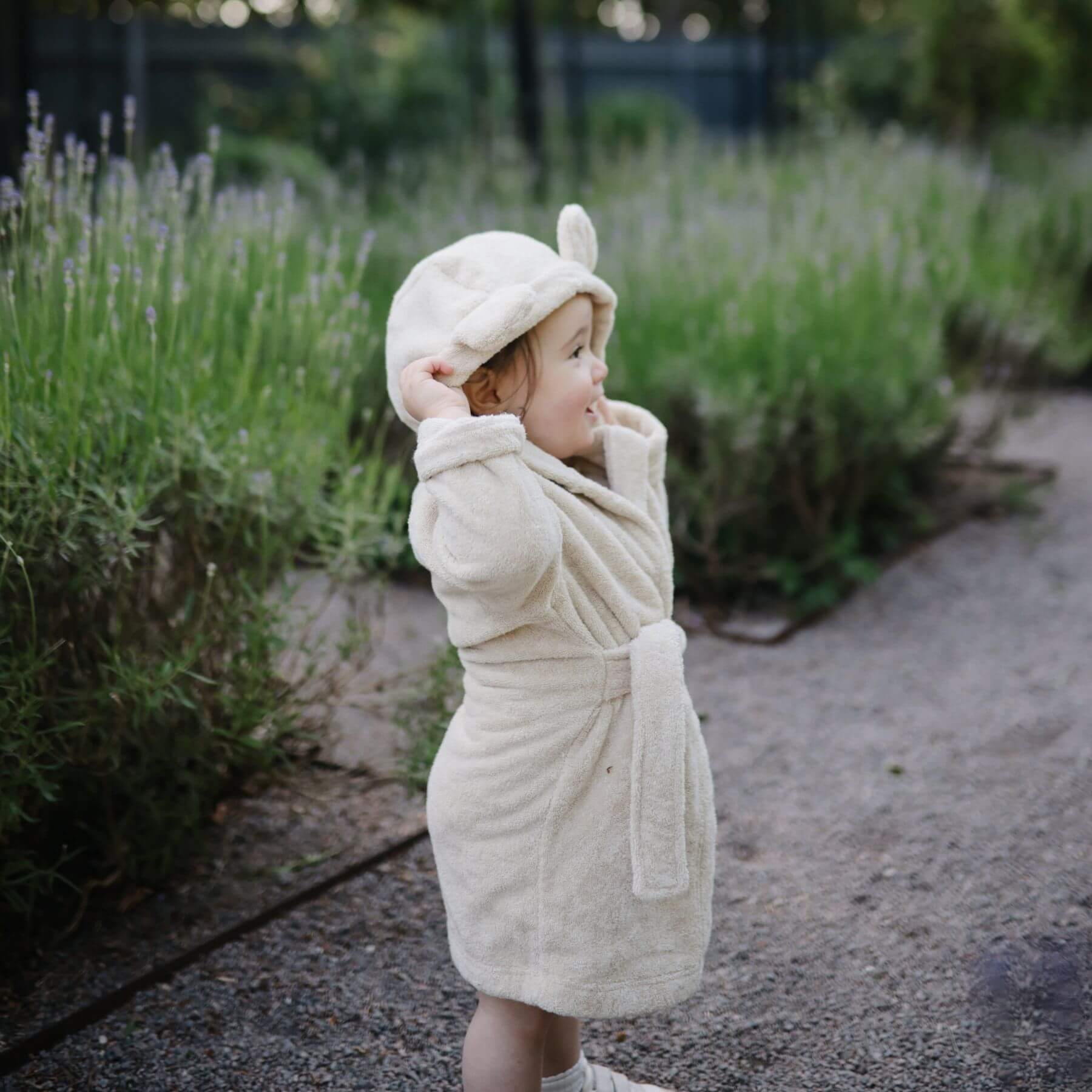 Child in Mushie Bear Robe standing in a garden with lavender plants.