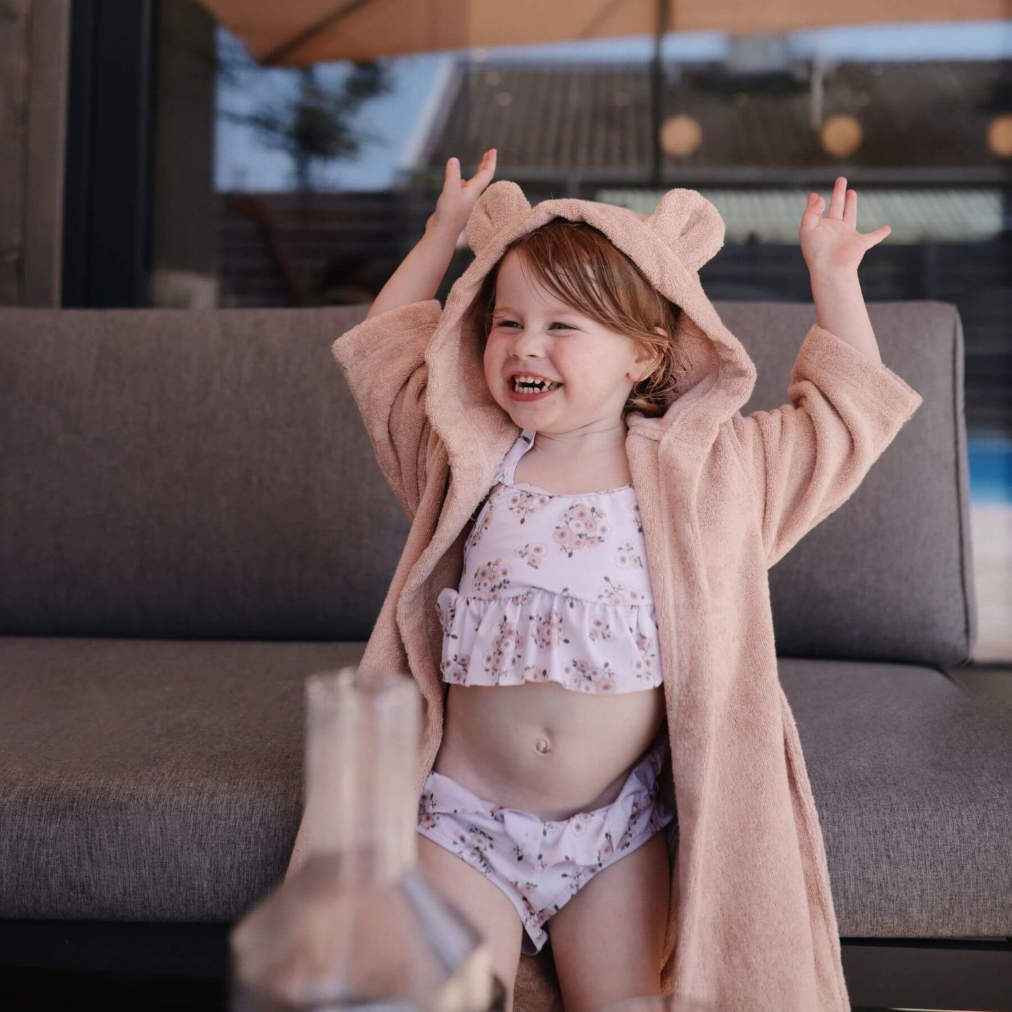 Child wearing a floral swimsuit and Mushie Bear Robe, standing on a couch.