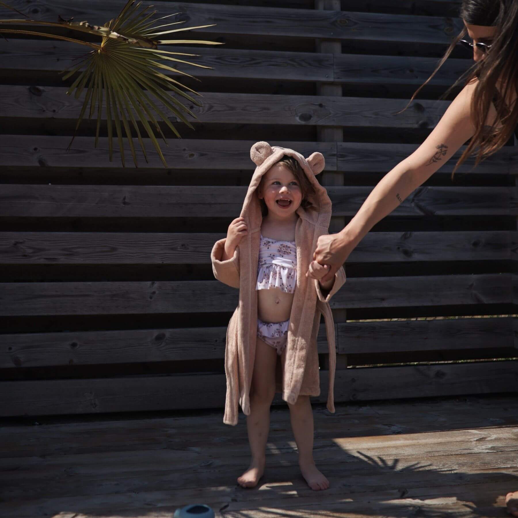 Child in Mushie Bear Robe standing next to an adult against a wooden background