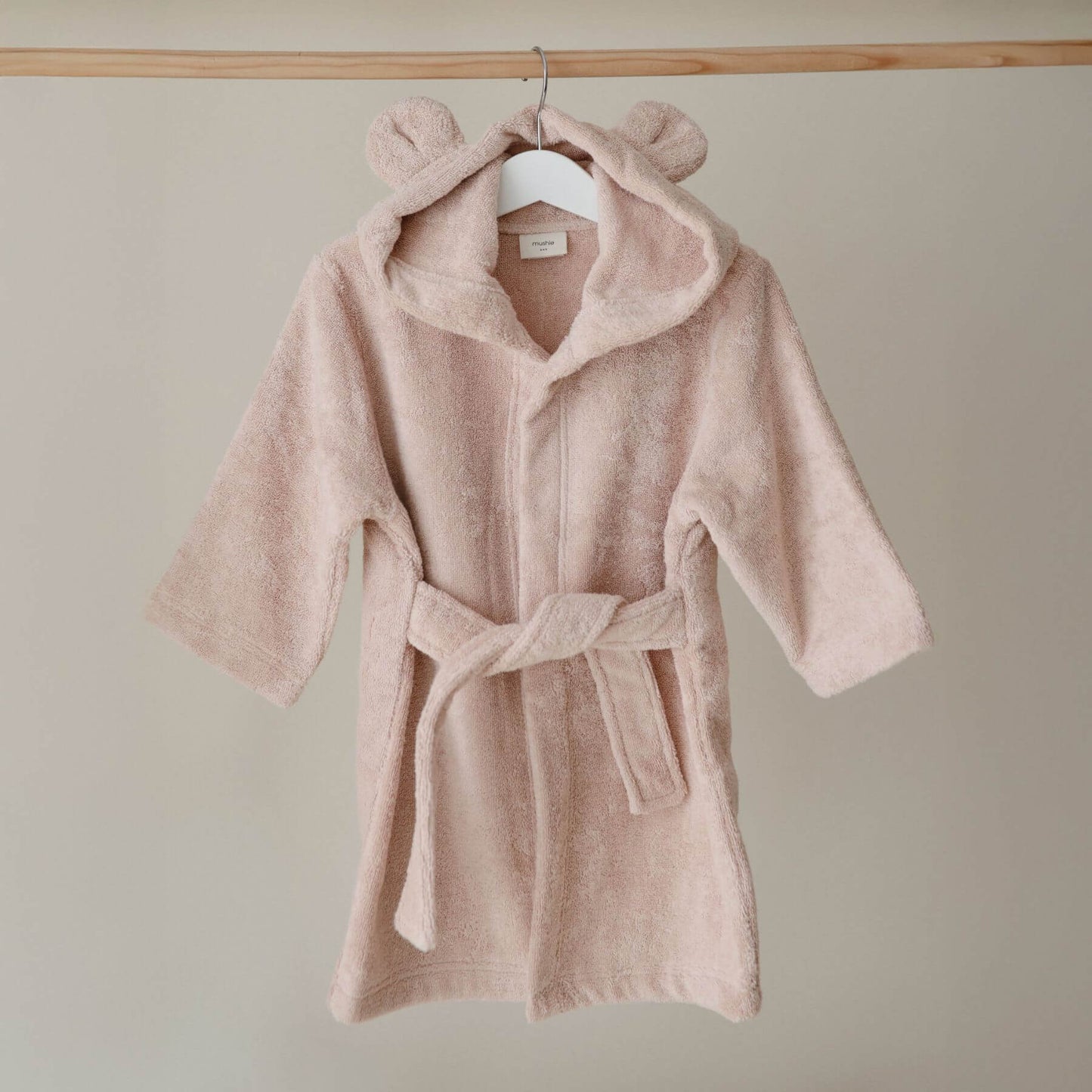 Mushie Bear Robe on a hanger against a beige wall.